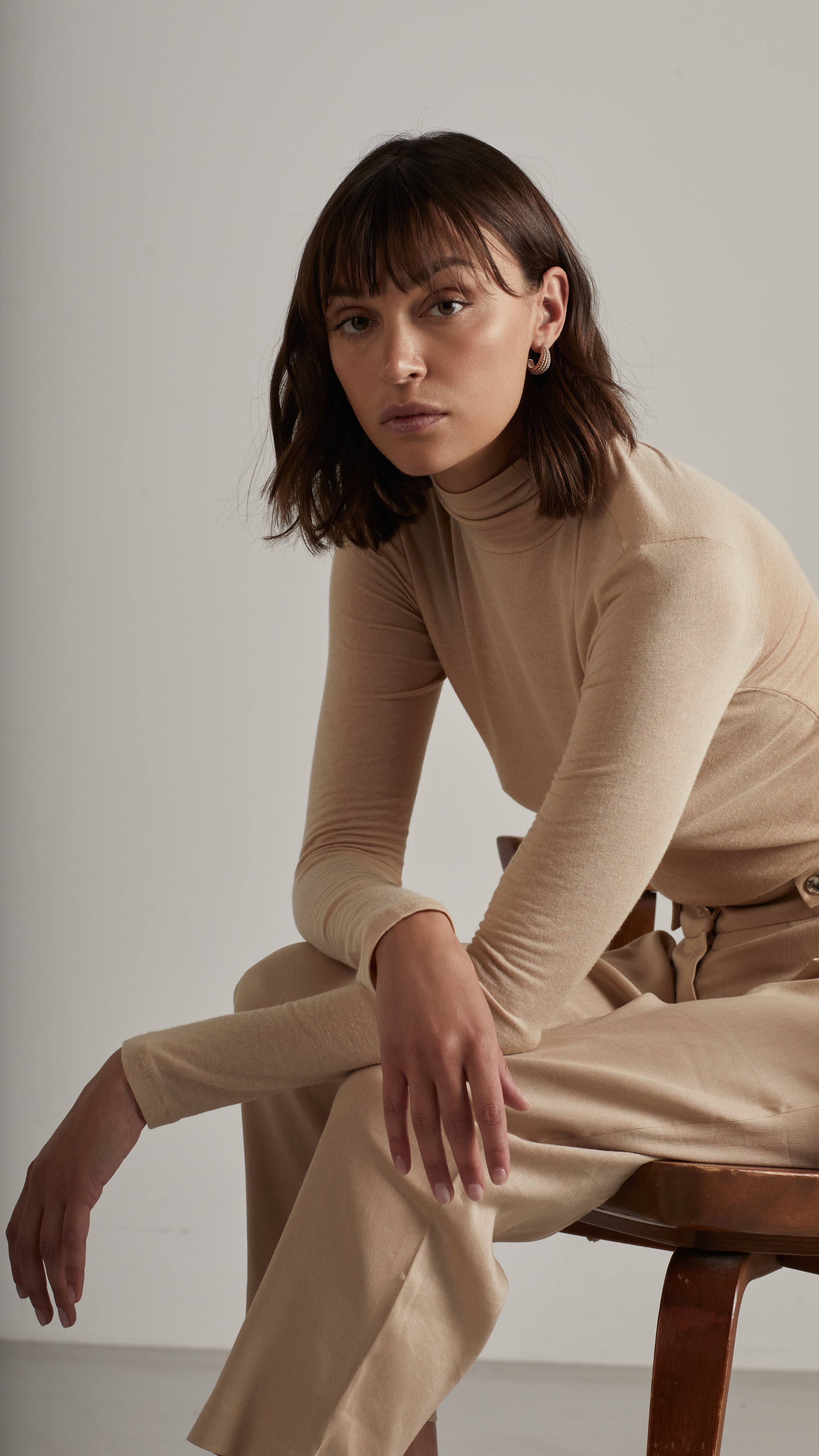 Woman in beige turtleneck and pants sitting on a wooden chair in a minimalist setting.