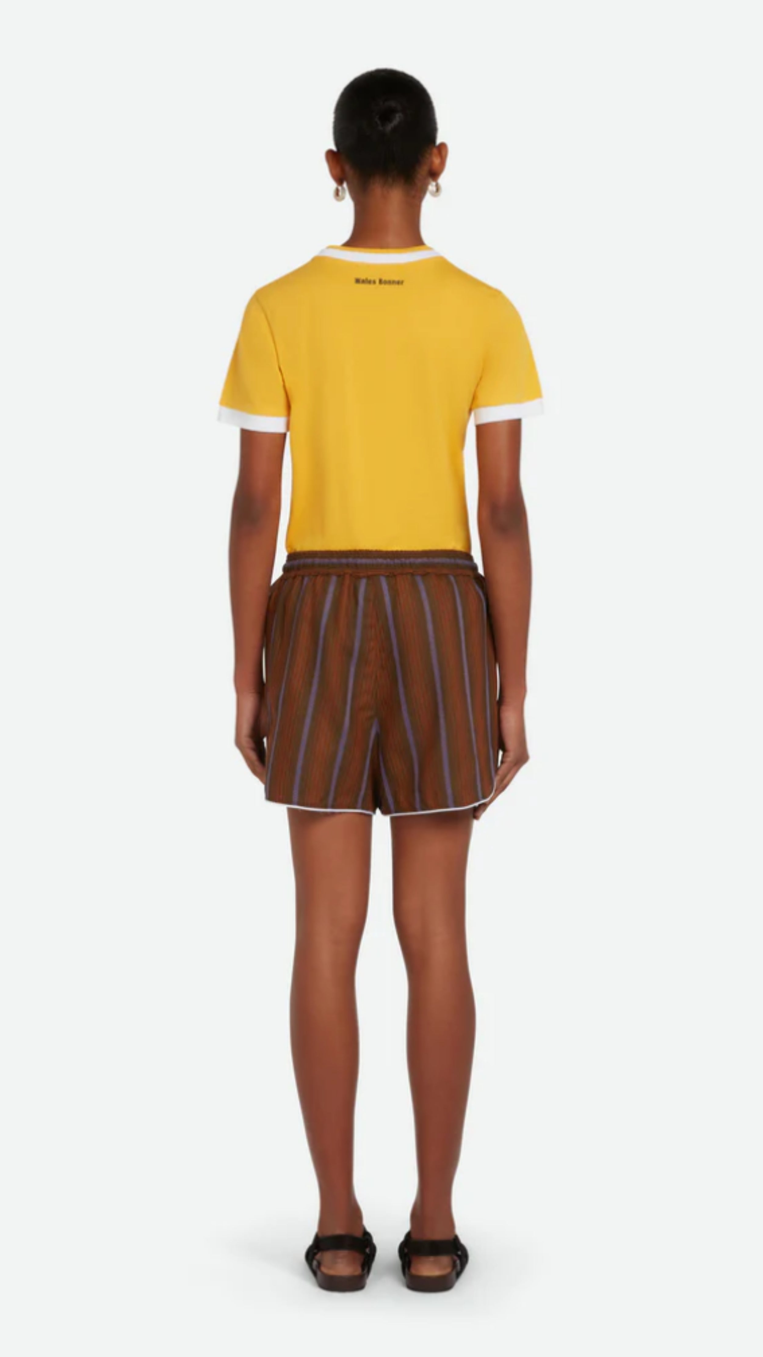 Wales Bonner The Life Shorts are made from Italian-made, lightweight herringbone summer wool fabric in deep brown and blue stripes. Classic atheltic shape these shorts have an elastic waistband, a draw string and contrasting white trim at the leg. Shown on model facing back.