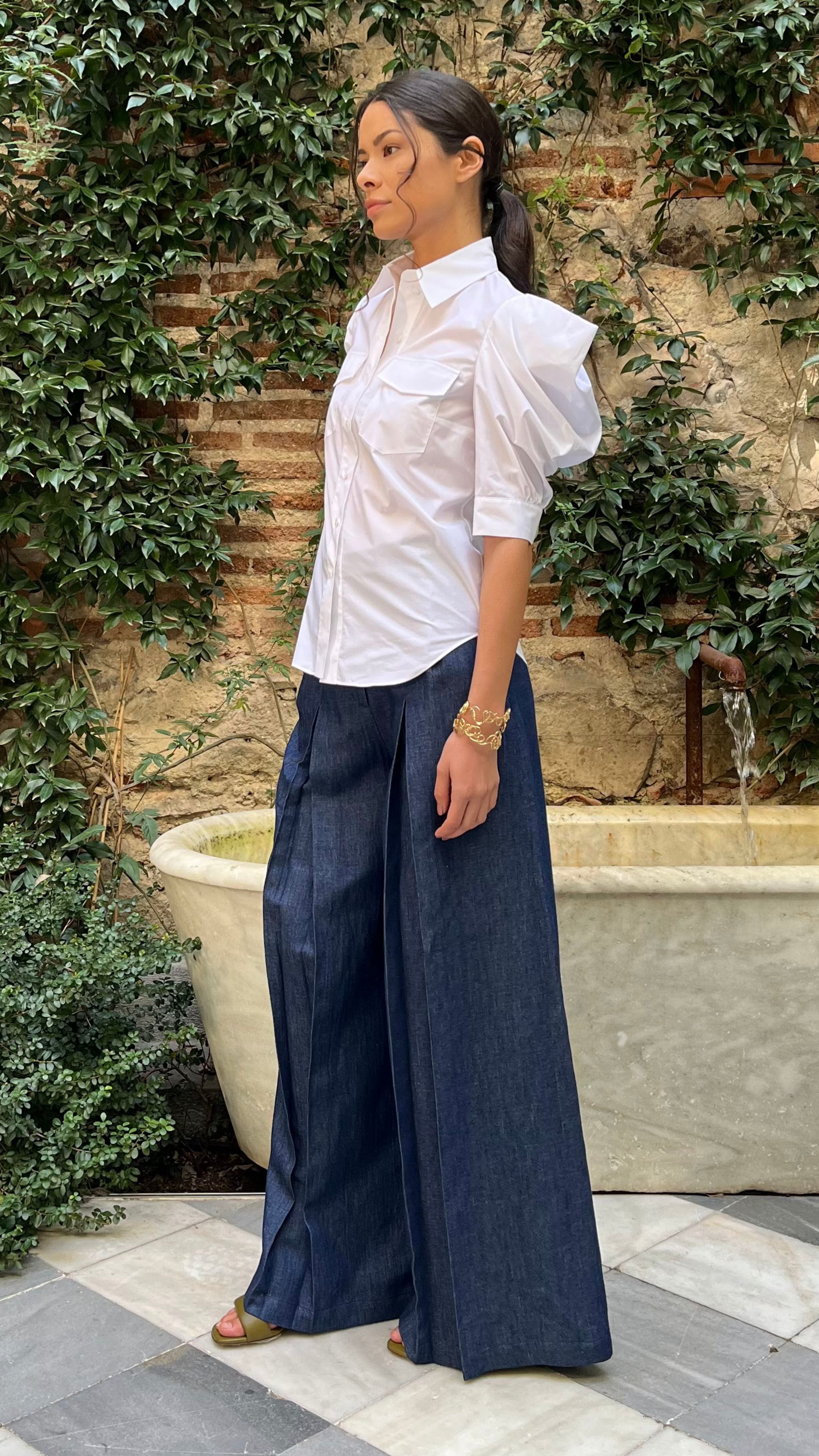 Rochas Paris Cotton Poplin Short Sleeved Shirt. A button up blouse in lightweight cotton it has two front pockets and puffed sleeves. Photo shown on model with a side view also wearing Rochas wide leg denim trousers.
