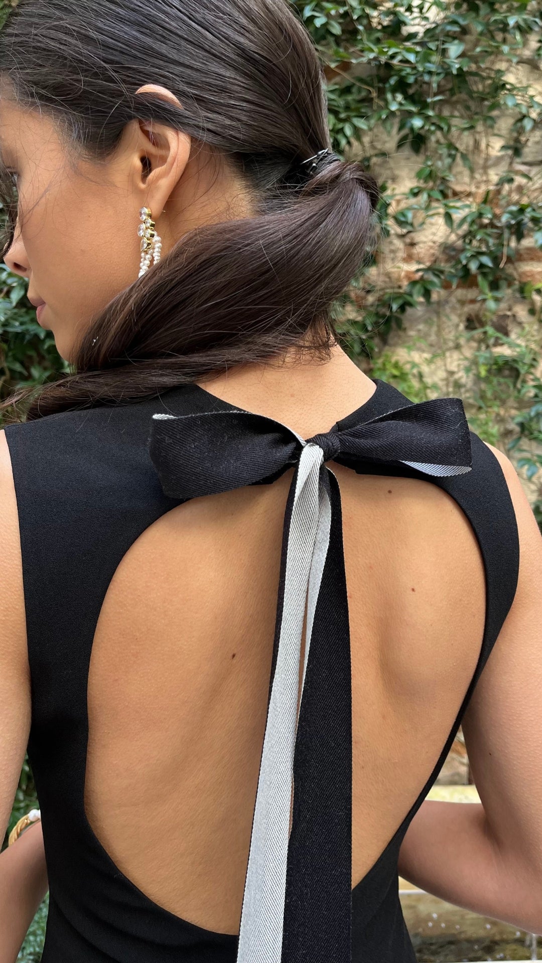 Roksanda Datura Dress. Black midi dress is crepe with front cut our panel and a line draped skirt. Midi length dress. Sleeveless. Open back with elegant tied bow at back nape neck. Shown on model close up of the back detail.