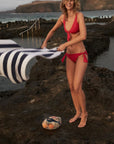 Talia Collins The Bow Tie Brief in Goji Red. Sustainable swimwear made from recycled materials. Classic side tie style with full coverage. California surfer chic. Experience 27 Madrid. shown on model at the beach.