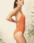 Talia Collins Cross Back Swimsuit in Terracotta Orange. Sustainable swimwear made from recycled materials. One piece classic swimsuit style with cross straps in the back. Scoop neck with full coverage bikini bathing suit top and bum coverage. Experience 27 Madrid. Shown on model facing side.