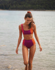 Talia Collins The Tricolor Athletic Top in Red, plum and orange. Sustainable swimwear made from recycled materials. Sports bra style bikini top. Scoop neck with full coverage bikini bathing suit top. Color block. Experience 27 Madrid. Shown on model at the beach.