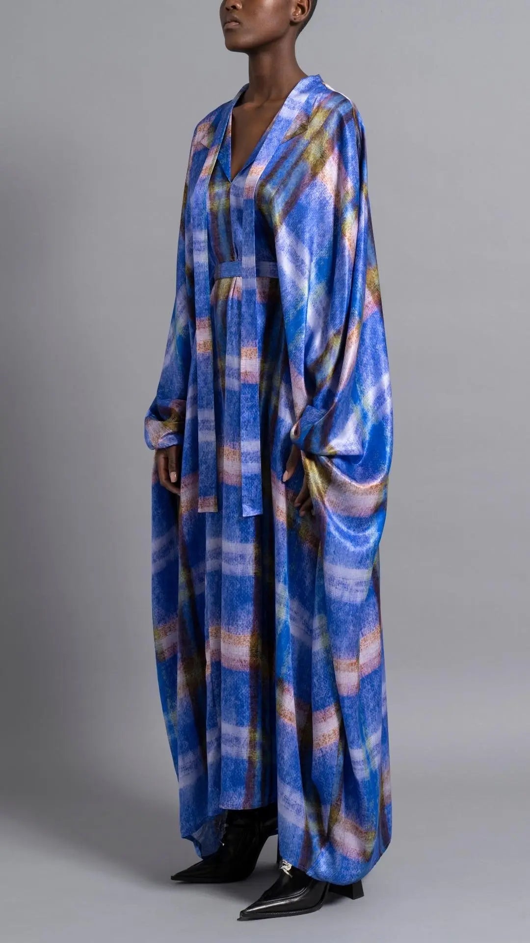 Thebe Magugu Blue Print Belted Kaftan. An elegant draped kaftan dress in blue with soft pink and grey striping. It features a V neck and can be belted to find the perfect fit for your body. It has long sleeves and is floor length. This photo shows the piece on the model facing to the front and side..