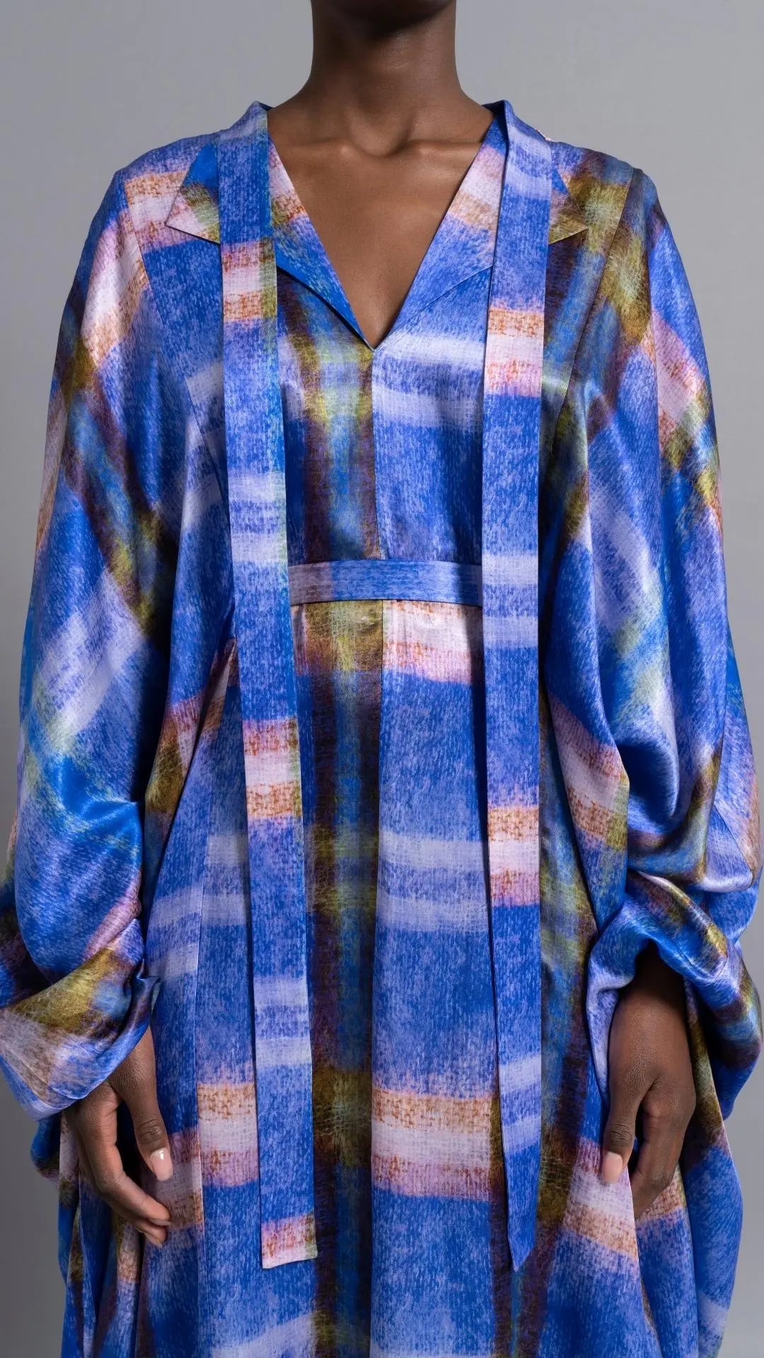 Thebe Magugu Blue Print Belted Kaftan. An elegant draped kaftan dress in blue with soft pink and grey striping. It features a V neck and can be belted to find the perfect fit for your body. It has long sleeves and is floor length. This photo shows a close up of the front top of the piece on the model facing front.