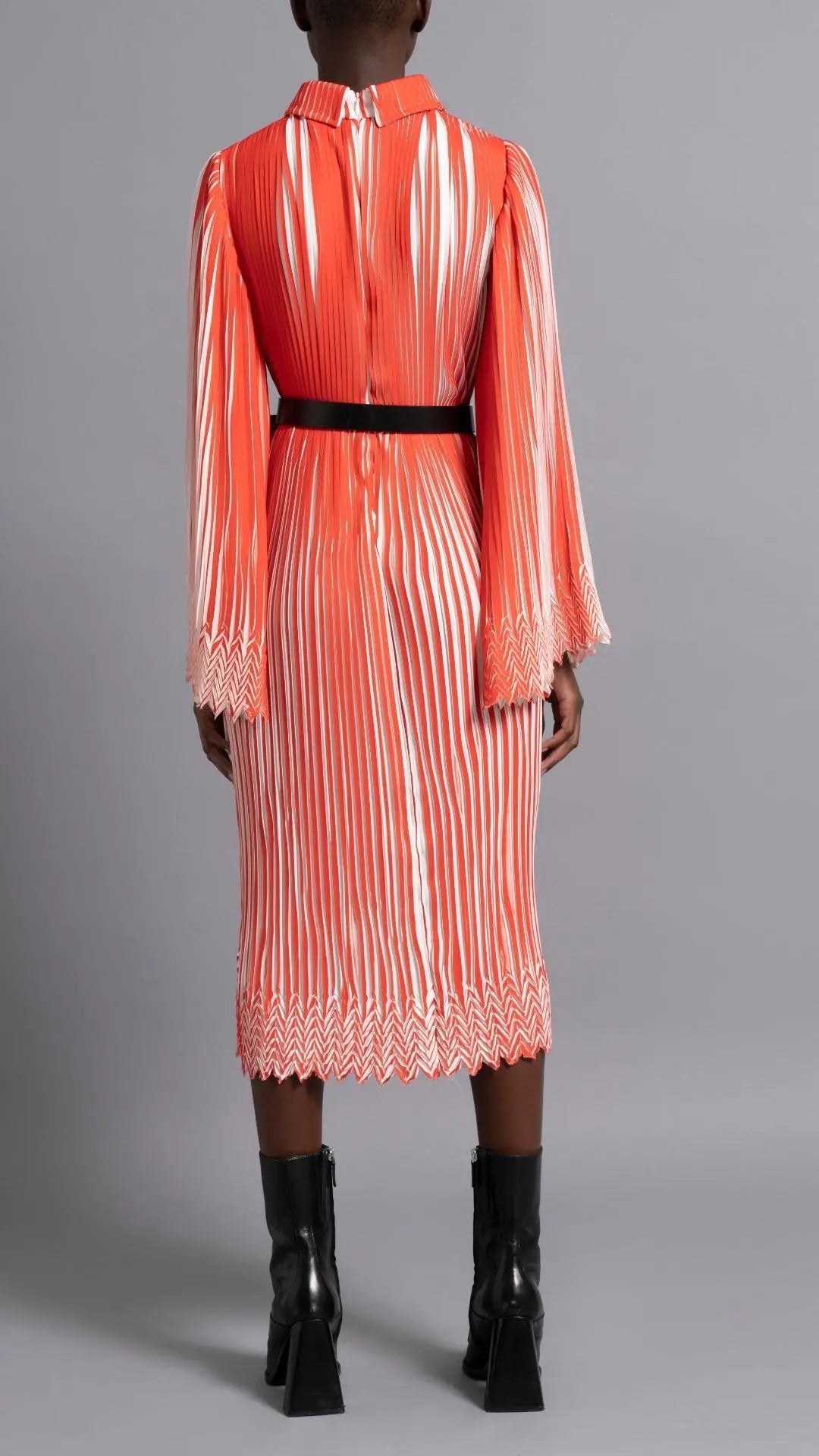 Thebe Magugu Chevron Pleated Dress. Stunning tomato red and white pleated dress with a collar, fitted waist and double chevron hem at the sleeves. It features a hidden zipper in the back. Shown on model facing back.