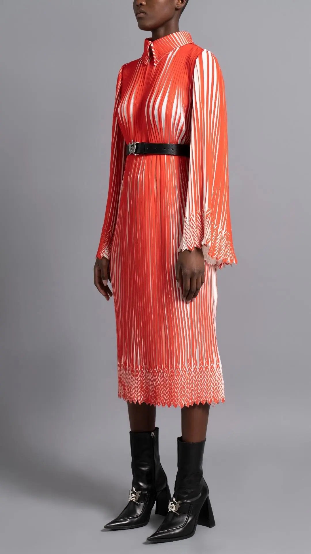 Thebe Magugu Chevron Pleated Dress. Stunning tomato red and white pleated dress with a collar, fitted waist and double chevron hem at the sleeves. It features a hidden zipper in the back. Shown on model facing to the side..