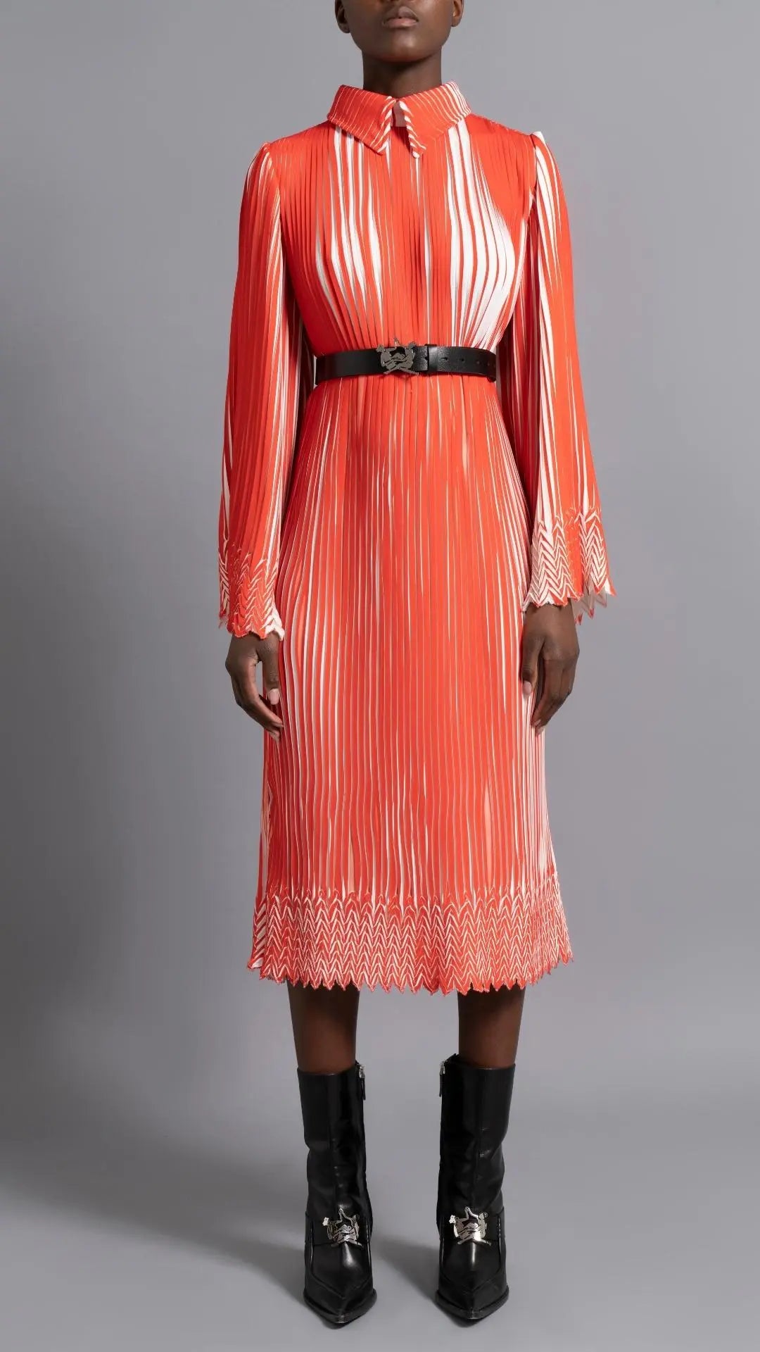 Thebe Magugu Chevron Pleated Dress. Stunning tomato red and white pleated dress with a collar, fitted waist and double chevron hem at the sleeves. It features a hidden zipper in the back. Shown on model facing forward.