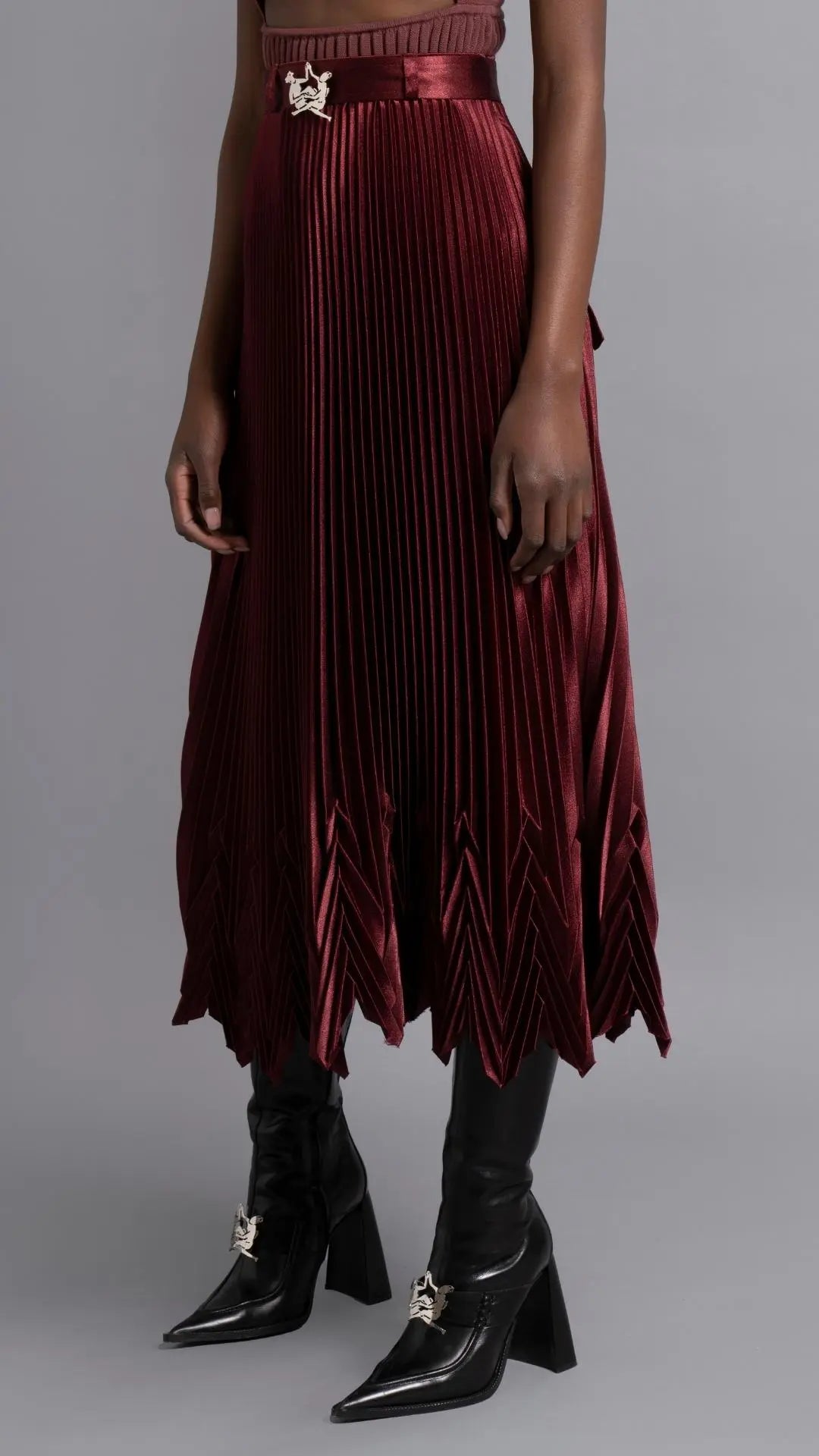 Thebe Magugu Chevron Pleated Skirt in a rich burgundy color. Fitted at the waist with straight pleats it falls into a double chevron pleat bottom. Long in length. It comes with a tie belt. Shown on model facing to the sise.