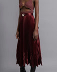 Thebe Magugu Chevron Pleated Skirt in a rich burgundy color. Fitted at the waist with straight pleats it falls into a double chevron pleat bottom. Long in length. It comes with a tie belt. Shown on model full view facing to the side..