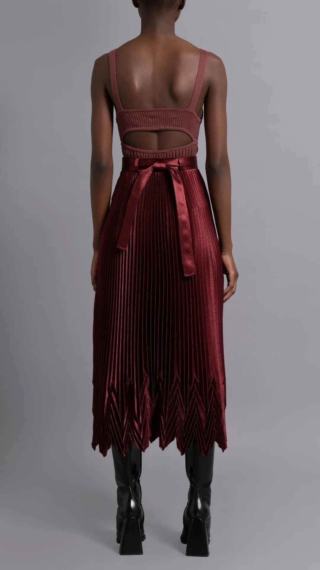 Thebe Magugu Chevron Pleated Skirt in a rich burgundy color. Fitted at the waist with straight pleats it falls into a double chevron pleat bottom. Long in length. It comes with a tie belt. Shown on model facing back.