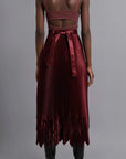 Thebe Magugu Chevron Pleated Skirt in a rich burgundy color. Fitted at the waist with straight pleats it falls into a double chevron pleat bottom. Long in length. It comes with a tie belt. Shown on model facing back.